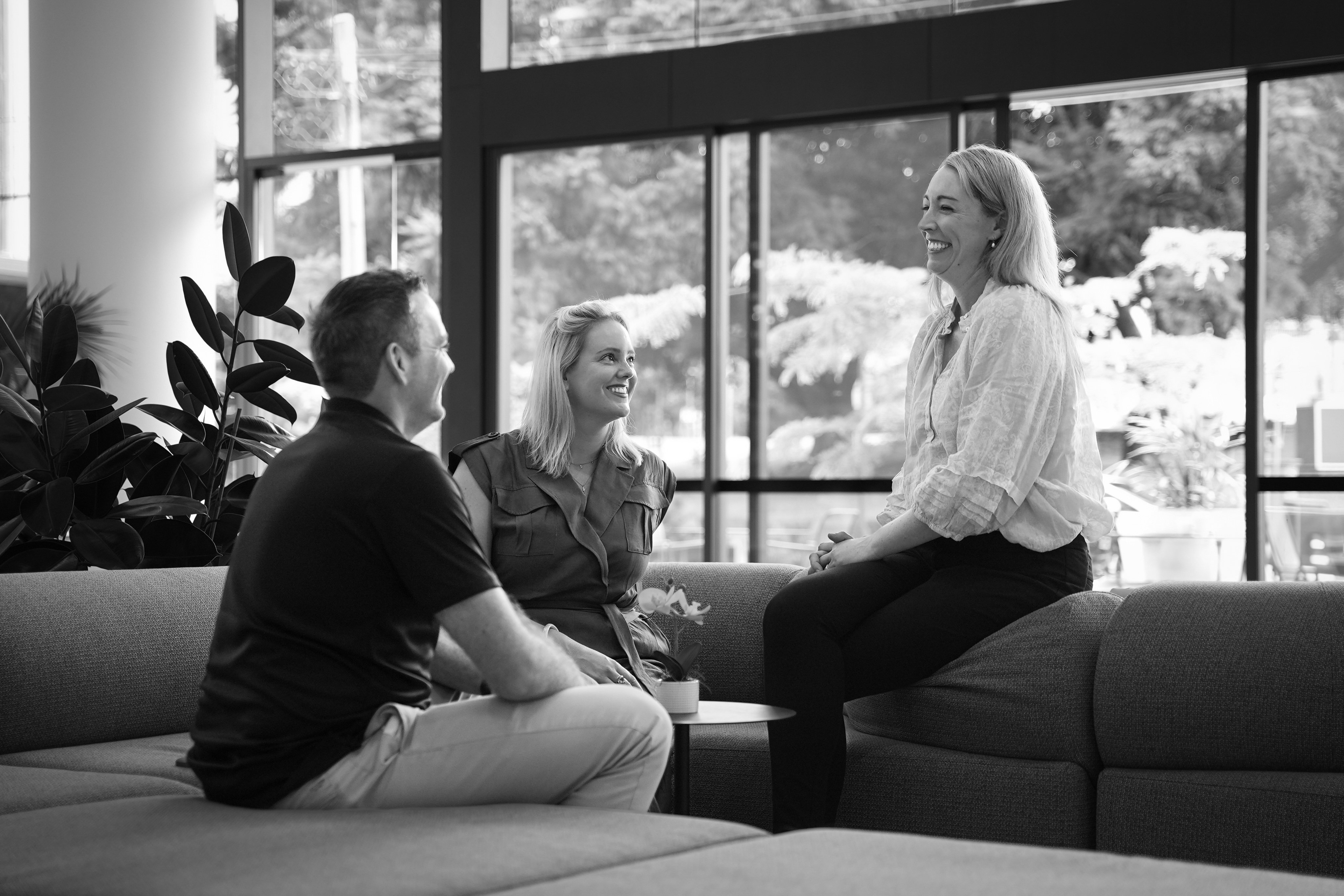 Three people smiling and talking while sitting on a modern sectional sofa in a bright office lobby.